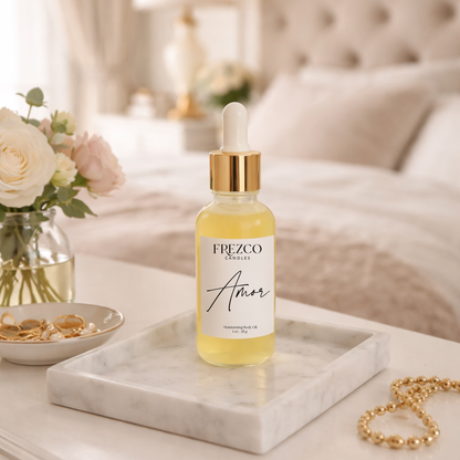 Frezco Candles 'Amor' body oil dropper bottle on a marble tray with flowers and jewelry in the background.
