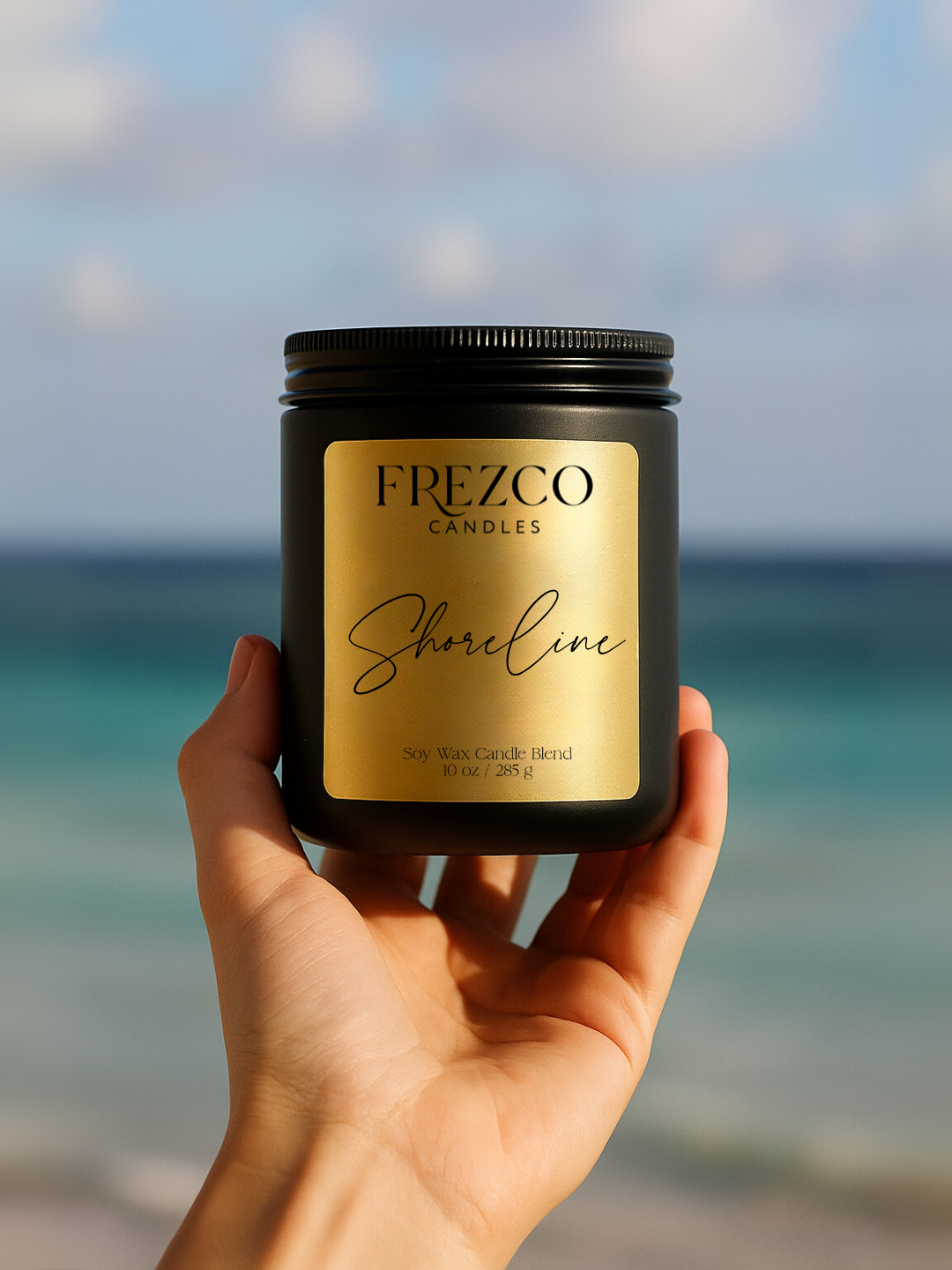 Hand holding a Frezco Candles 'Shoreline' candle with a beach background