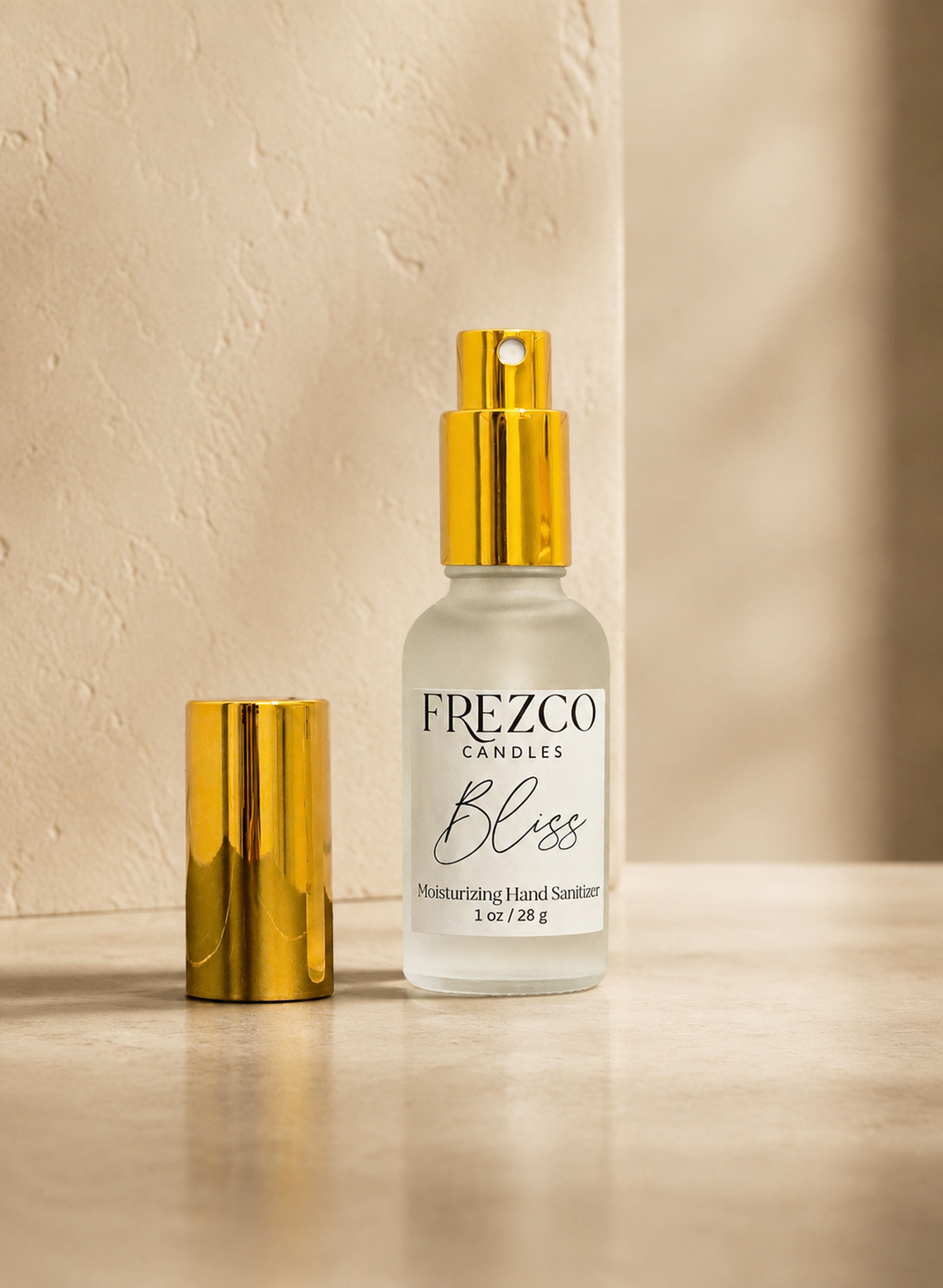 A bottle of Frezco Candles 'Bliss' moisturizing hand sanitizer with a gold spray top, shown next to its gold cap on a beige stone surface against a textured beige wall.