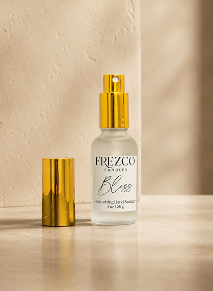 A bottle of Frezco Candles 'Bliss' moisturizing hand sanitizer with a gold spray top, shown next to its gold cap on a beige stone surface against a textured beige wall.