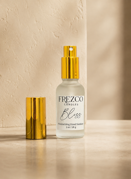 A bottle of Frezco Candles 'Bliss' moisturizing hand sanitizer with a gold spray top, shown next to its gold cap on a beige stone surface against a textured beige wall.