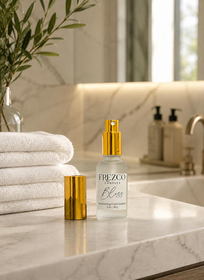 A spray of Frezco Candles 'Bliss' sanitizer spray with a gold spray cap and its matching gold cap placed beside it on a white marble bathroom counter, with folded white towels and a plant in the background.