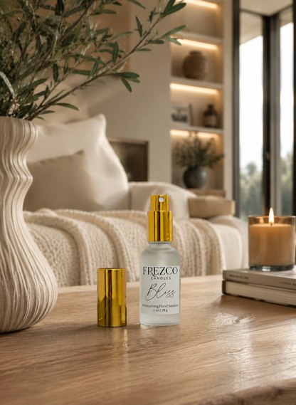 A clear glass sanitizer hand spray with a shiny gold pump and matching gold cap sits on a wooden table in a cozy, softly lit living room. The background shows a cream sofa, shelves with warm lighting, and a lit candle on books.