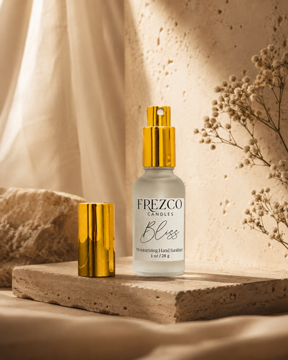 Frezco Candle Bliss moisturizing Hand Scent in a frosted glass bottle with a shiny gold pump, displayed next to its gold cap on a textured stone slab with warm, draped fabric and dried flowers in the background.