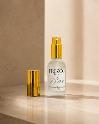 FREZCO Candles Bliss moisturizing hand sanitizer in a frosted glass bottle with a shiny gold pump, shown next to its detached gold cap on a beige stone surface.