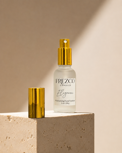 Frezco Candles 'Elégance' moisturizing hand sanitizer in a frosted glass bottle with a shiny gold pump, displayed next to its detached gold cap on a textured beige stone pedestal against a warm beige background.
